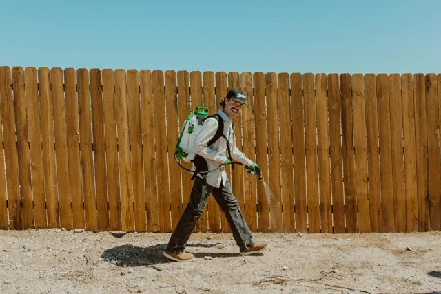 NEARPEST technician using a backpack sprayer along a wooden fence in a desert yard