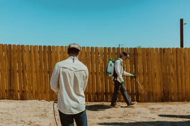 Two NEARPEST team members beside a wooden fence during perimeter treatment