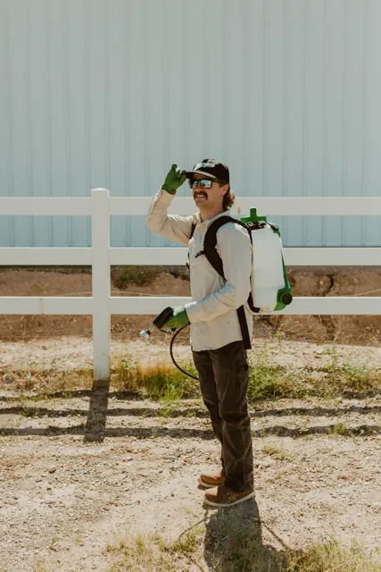 NEARPEST team member wearing a backpack sprayer beside a white rail fence