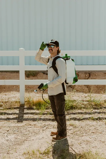 NEARPEST team member wearing a backpack sprayer beside a white rail fence