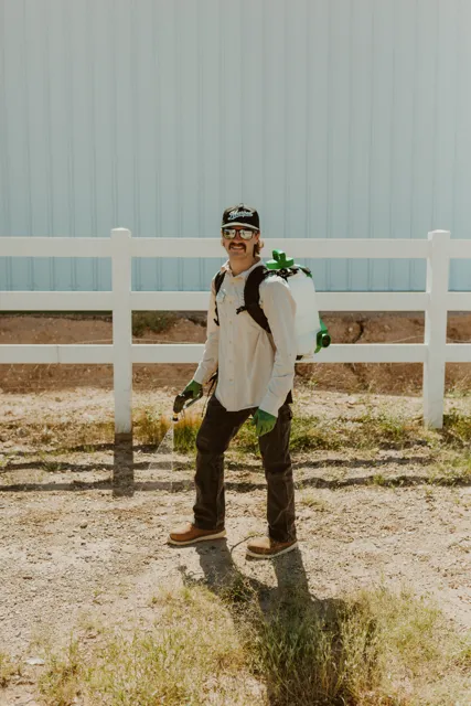 NEARPEST team member wearing a backpack sprayer beside a white rail fence