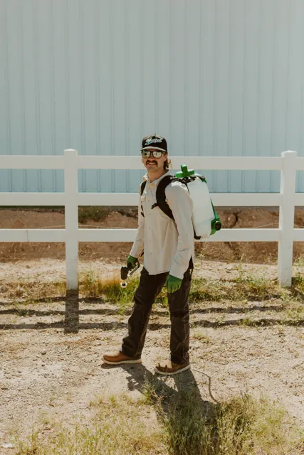 NEARPEST team member wearing a backpack sprayer beside a white rail fence