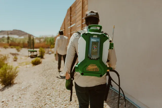 NEARPEST technician using a backpack sprayer beside a wooden fence in a desert yard