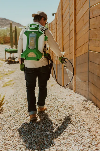 NEARPEST technician using a backpack sprayer beside a wooden fence in a desert yard
