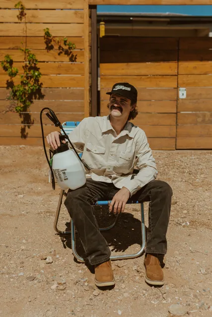 NEARPEST team member seated beside a green backpack sprayer outside a wood-sided building