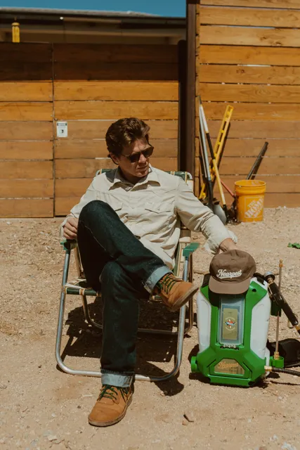 NEARPEST team member seated beside a green backpack sprayer outside a wood-sided building